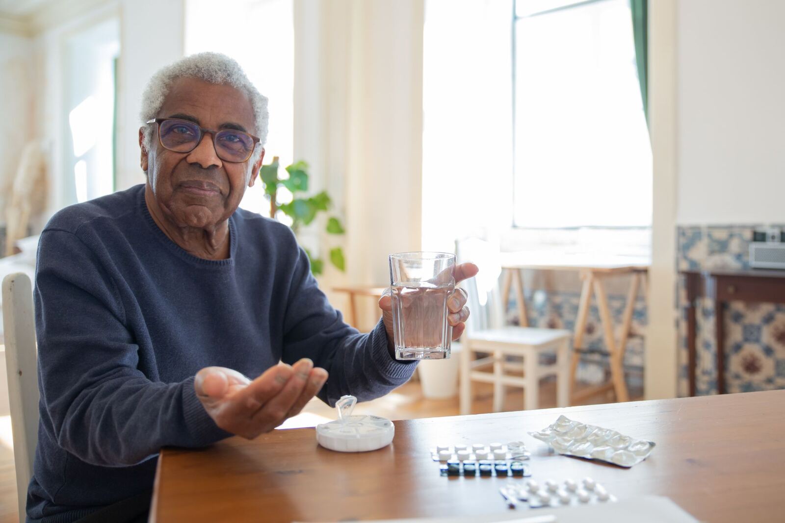 An older man at home with a glass of water and his daily medication on the table